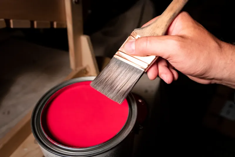 A brush is going to be dipped in a red paint can.