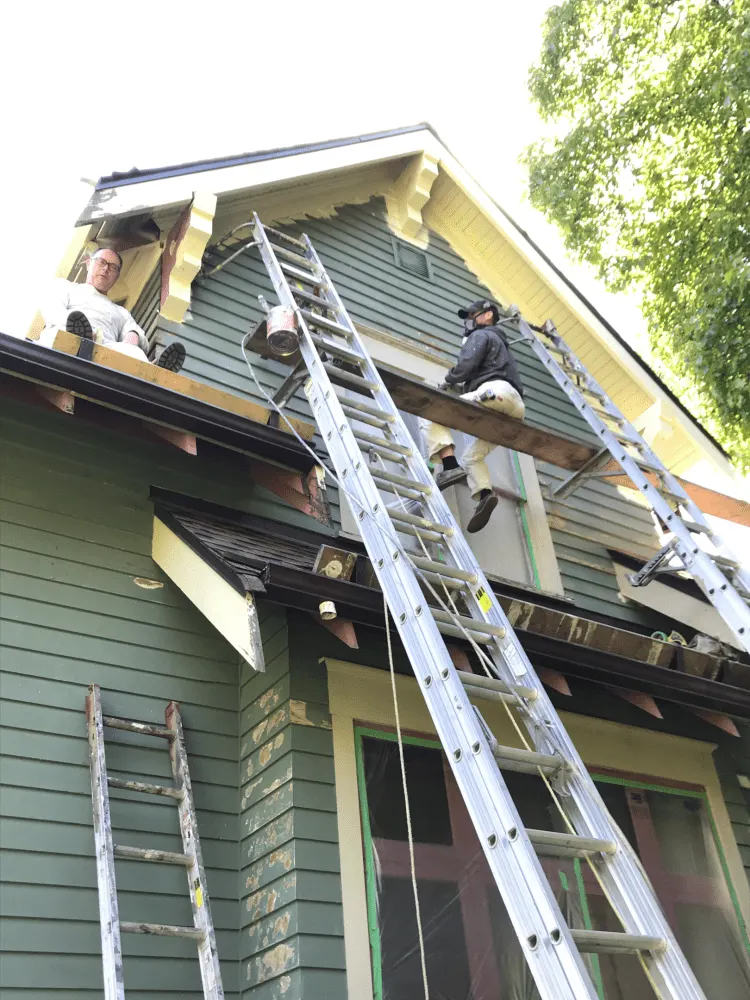 A painting contractor is painting house exterior wall in a dark cyan colour. And 3 ladders are leaning towards the the wall.