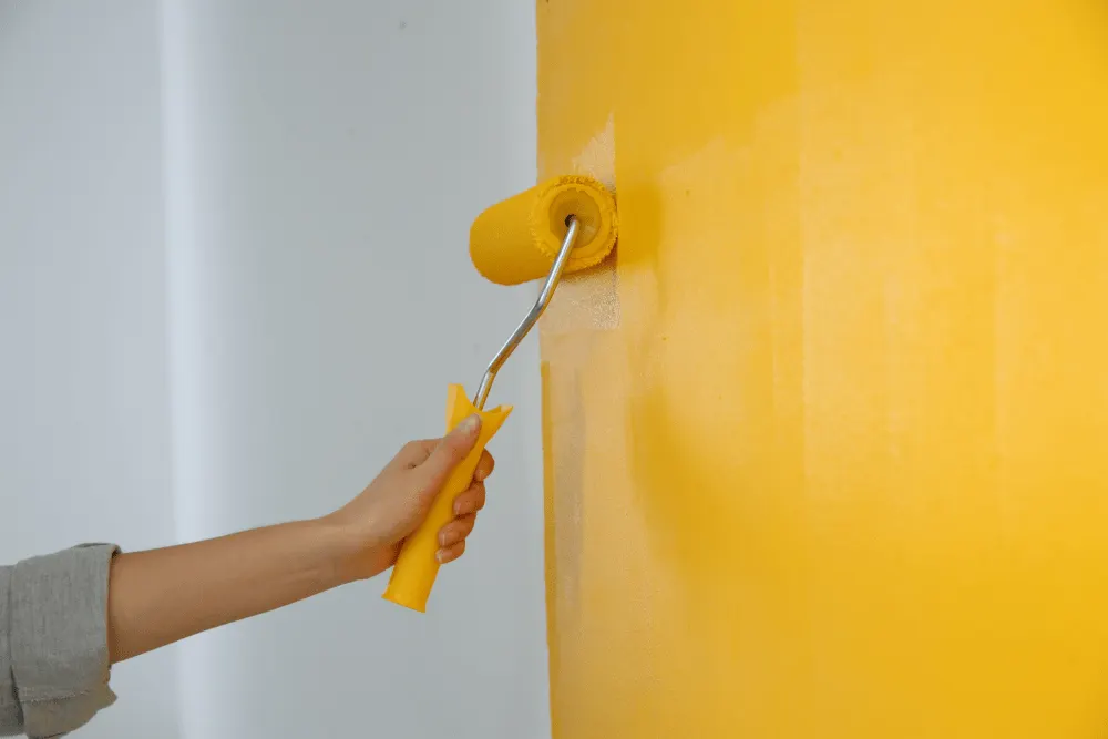A hand with painting roller is painting interior wall in yellow colour.