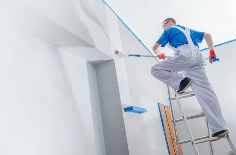 The painting contractor is painting an interior wall in a white colour.