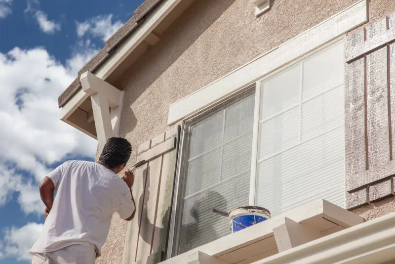 The painting contractor is painting a window frame in a light beige colour.
