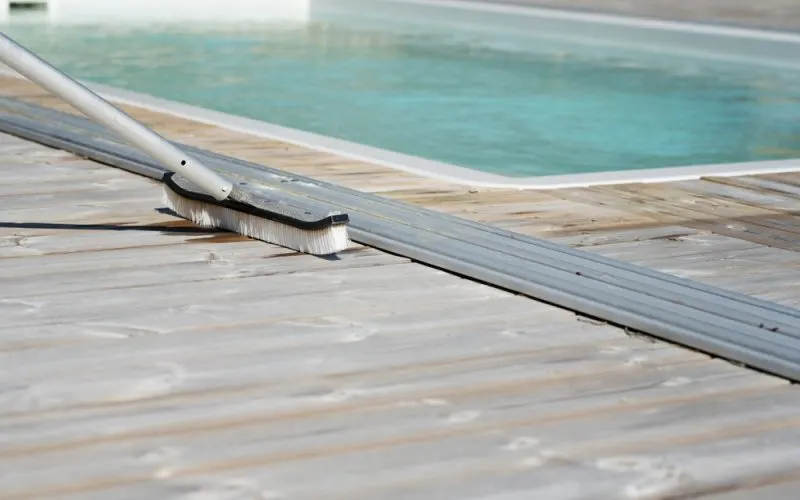 A wooden deck being stained, with a brush applying the stain and a clear pool in the background.