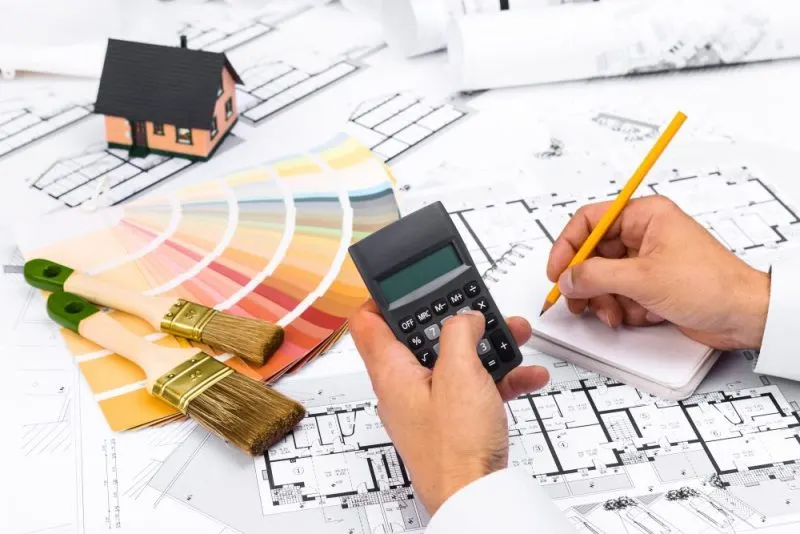 A man doing paint calculation with calculator, drawings, brushes, and a small house.