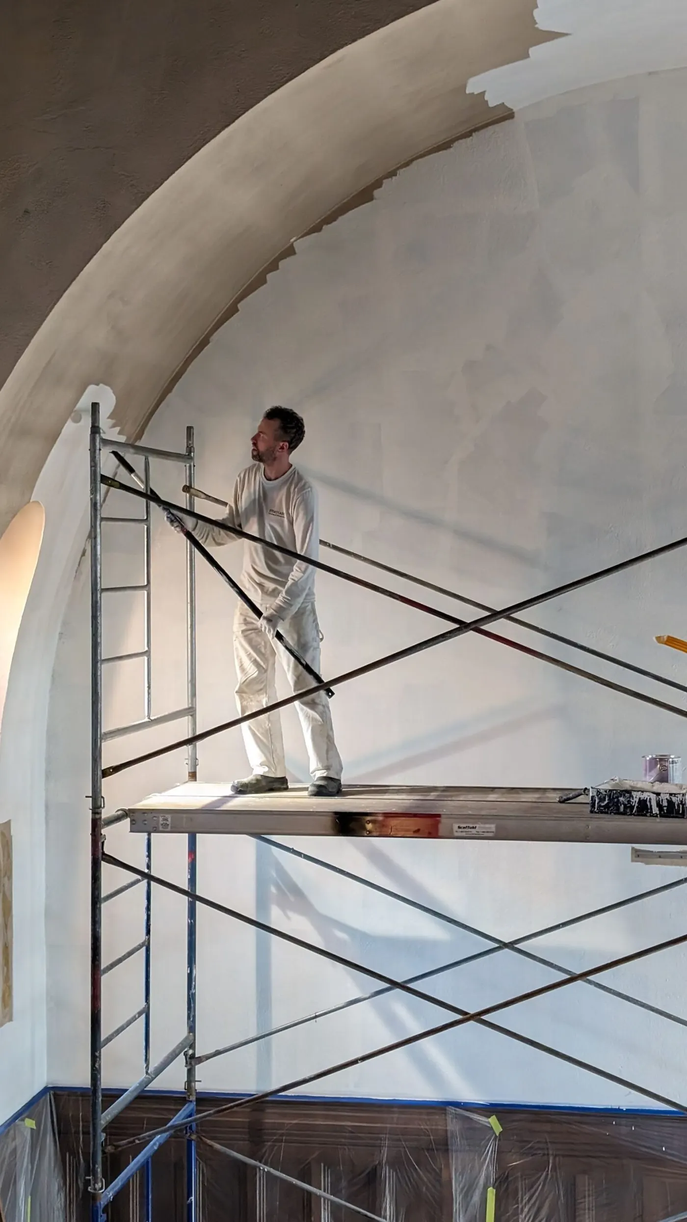 Painter on scaffolding applying white paint to a large curved interior wall with a roller at a commercial site.