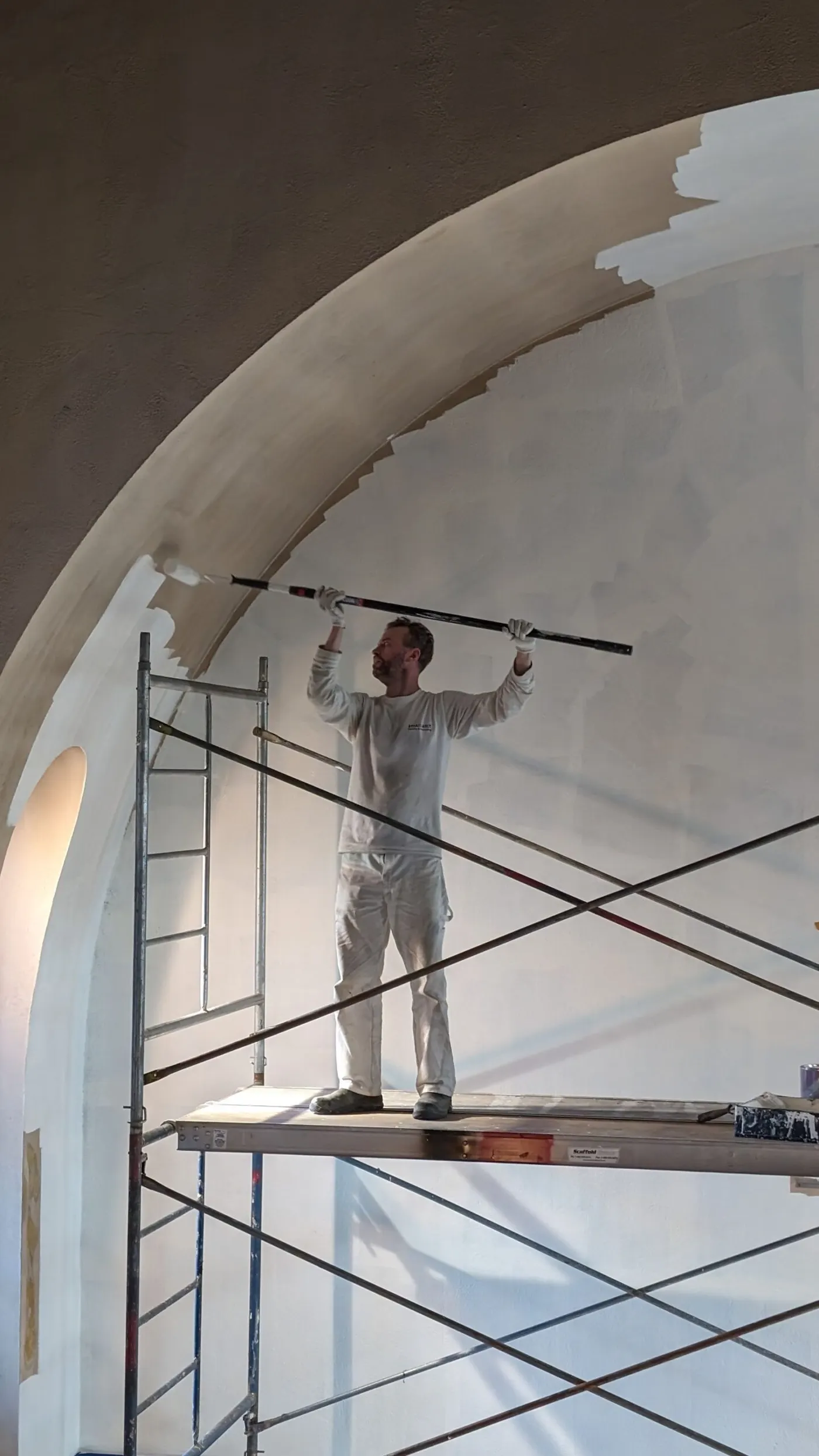 Painter on scaffold applying white paint to a tall arched wall in a strata building interior during repainting project.
