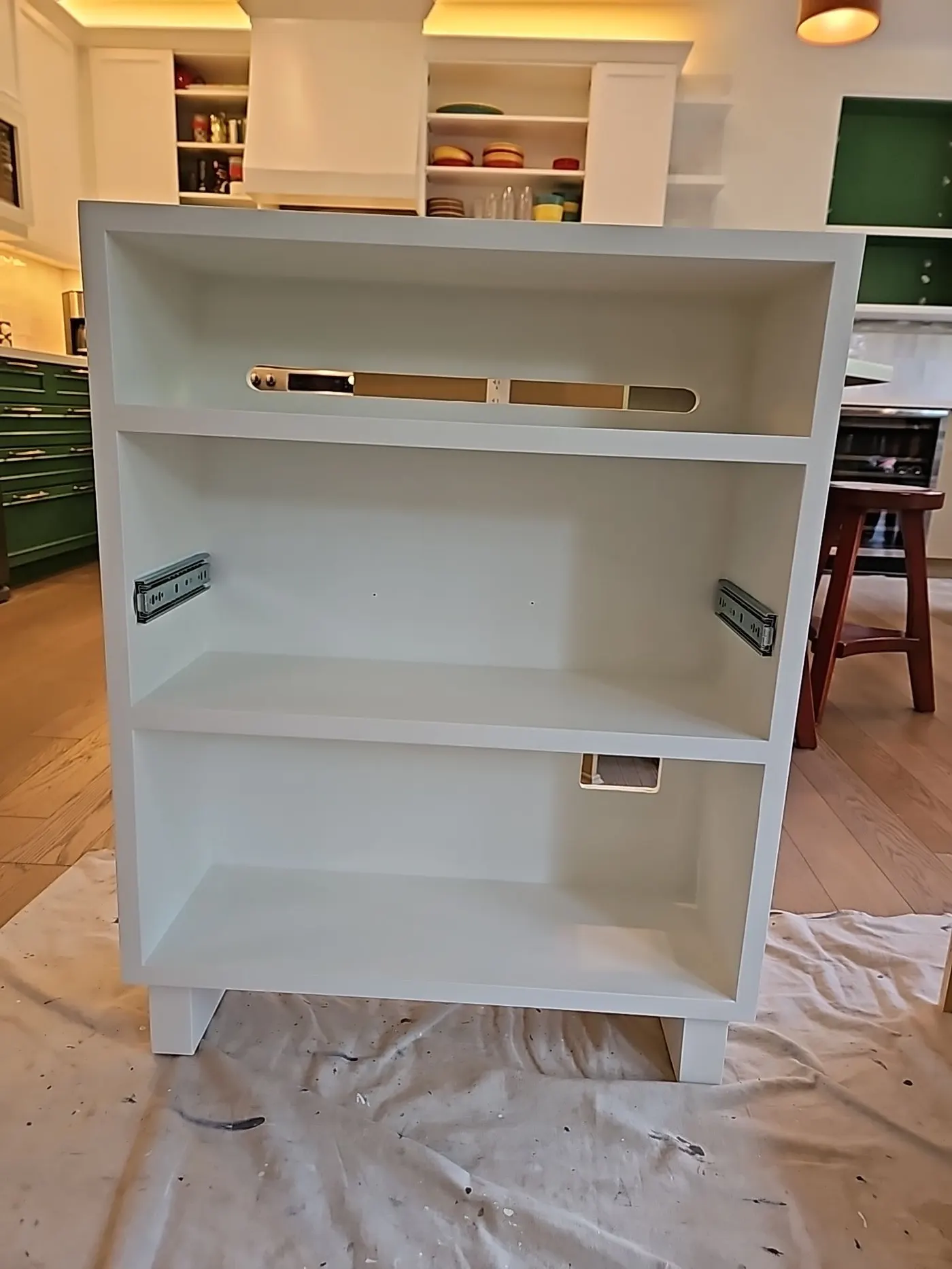 Newly painted white cabinet with open shelves and drawer slides, positioned on a drop cloth in a kitchen, showcasing precise paint work and preparation for installation.
