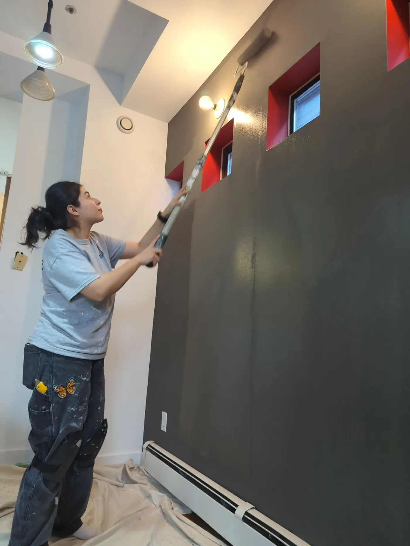 Woman painting an interior accent wall dark gray using a roller with extension pole; drop cloth protects floor below.