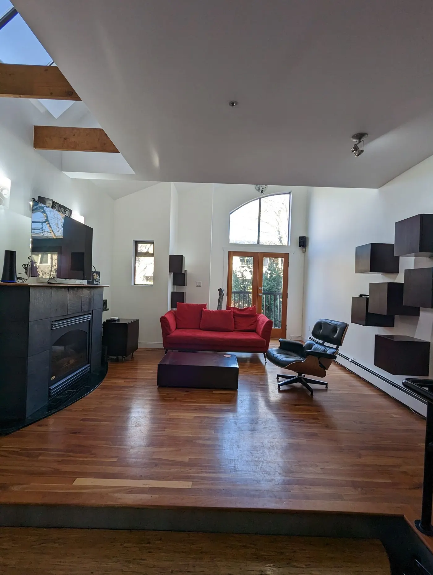Modern living room with red sofa, hardwood floors, white walls, and vaulted ceiling with exposed beams and skylight.