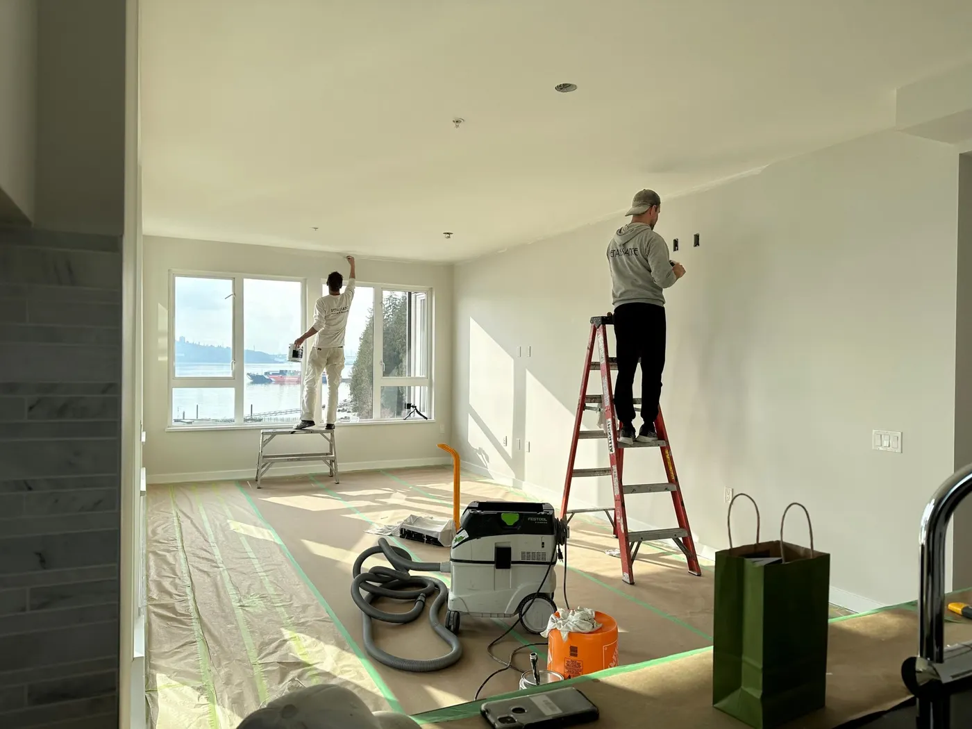 Two painters are painting the interior drywalls in a room white.