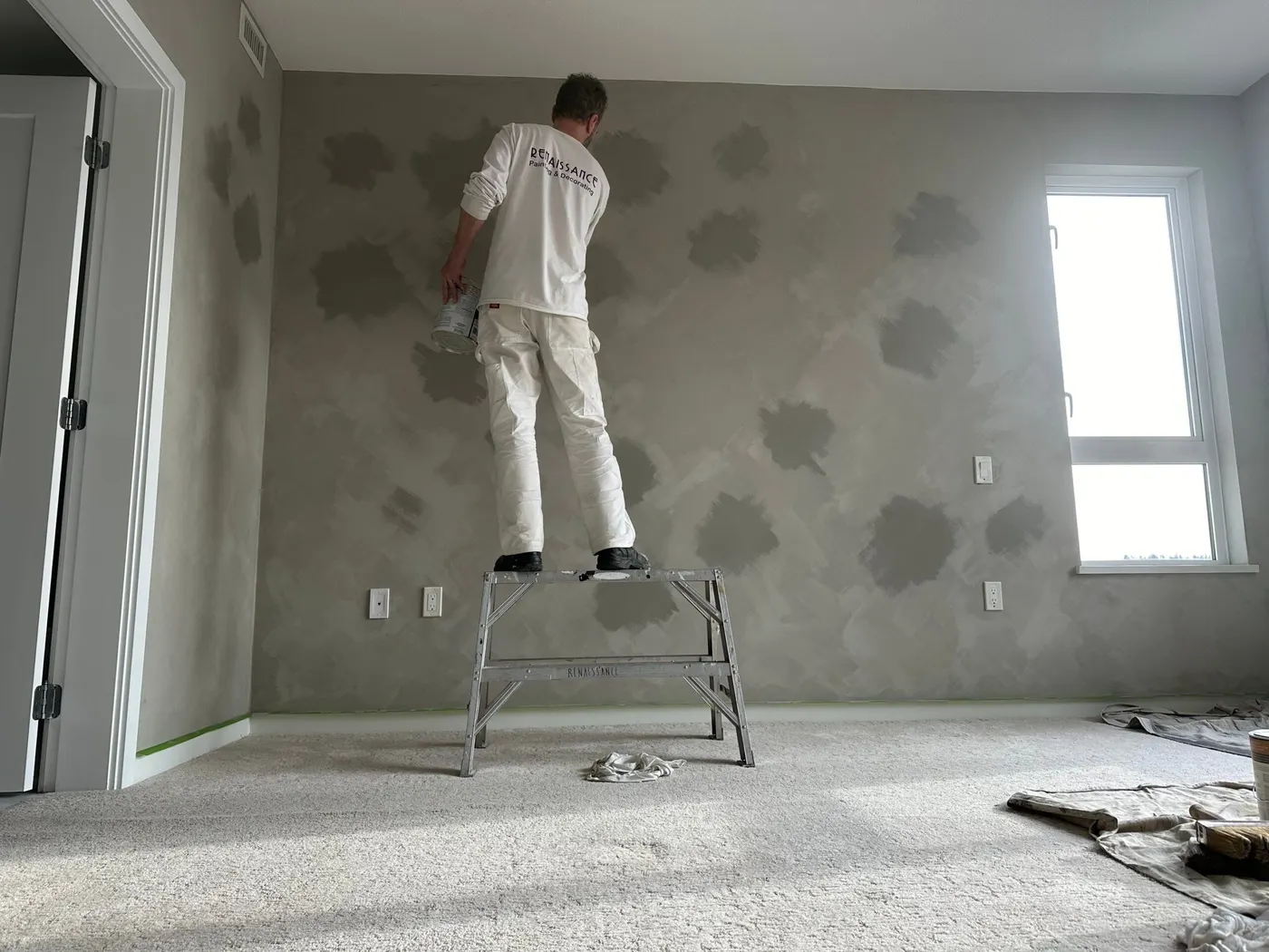 Painter is painting a drywall in a grey colour on a step ladder.