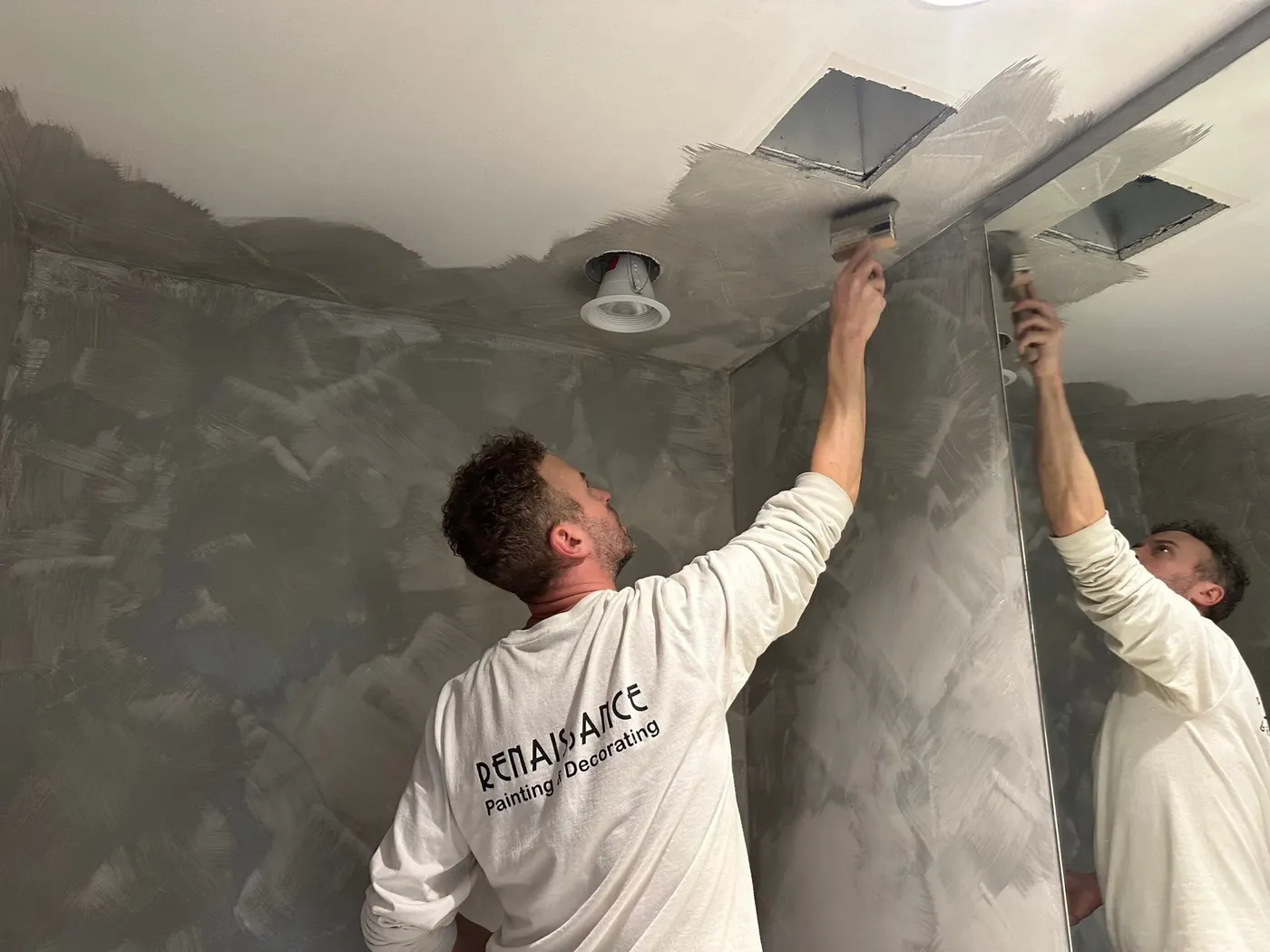 A painter is painting a celling in a grey colour in front of the mirror.