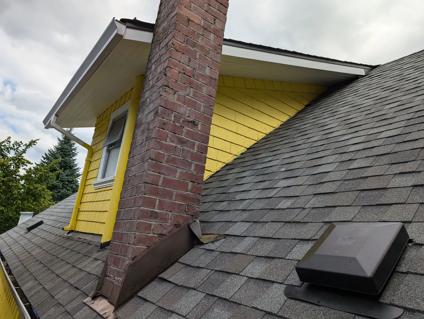 Brick chimney beside a freshly painted yellow dormer and gray asphalt shingle roof on a Vancouver, BC character home.