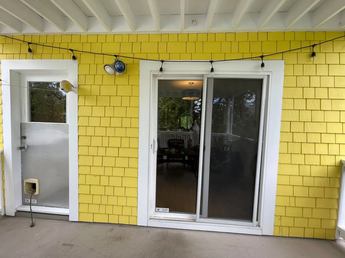 Completed exterior paint job showing bright yellow shingle siding, a white-trimmed sliding glass door, and string lights on a Vancouver home's back porch.