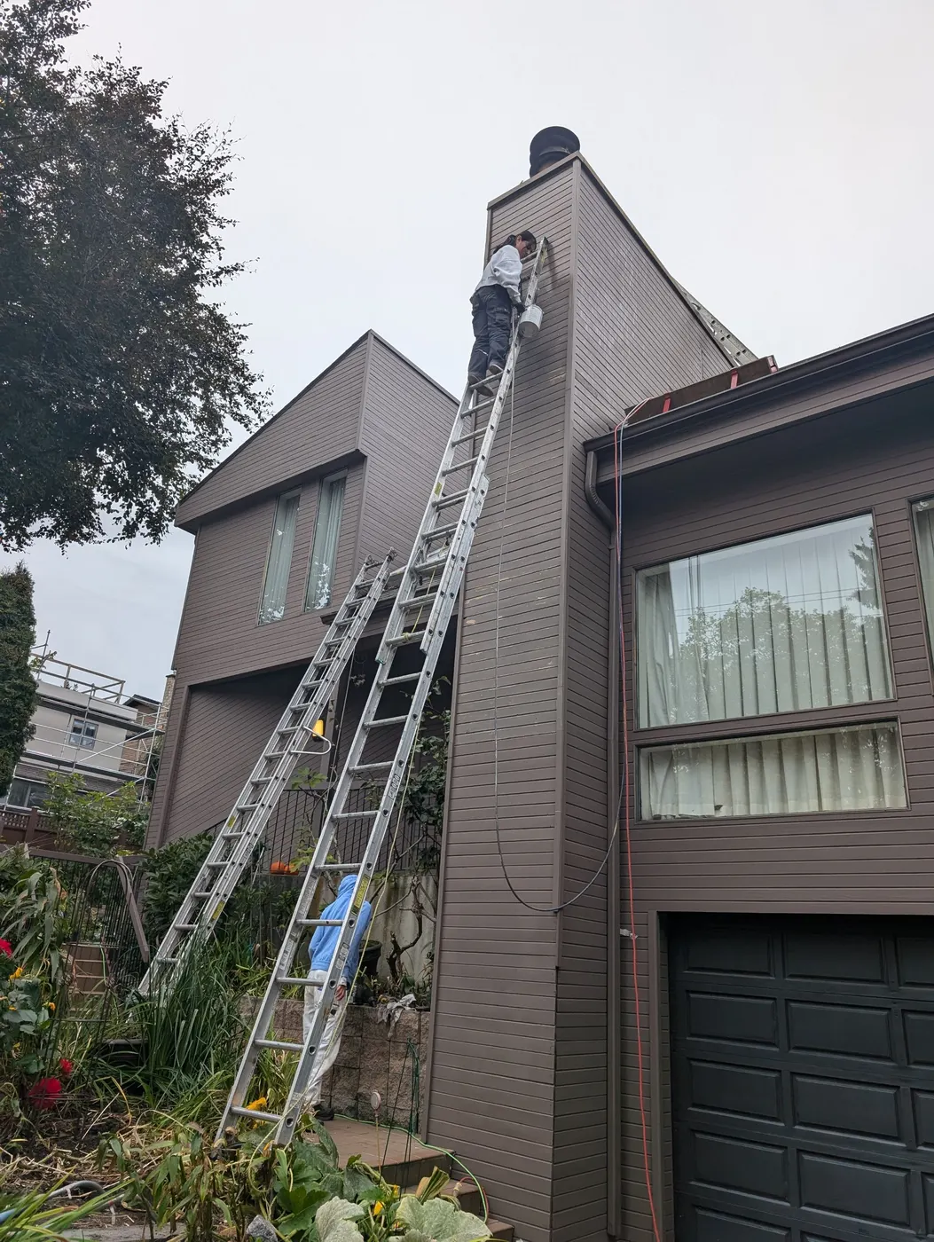 Painter high on a ladder applying primer to chimney siding on a Vancouver, BC residential exterior.
