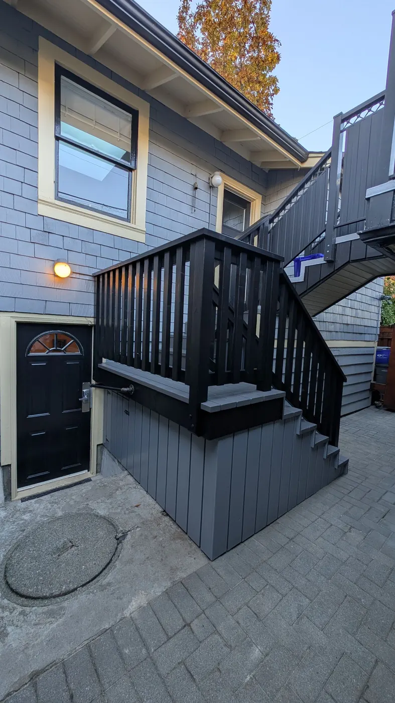 Fully rebuilt exterior wood staircase with black railings and warm porch lighting at dusk on a Vancouver, BC home