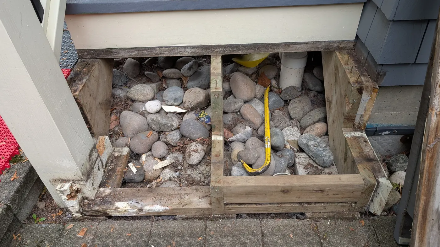 Close-up view of rotted wood framing removed from beneath an exterior staircase, exposing rock drainage bed, Vancouver BC.