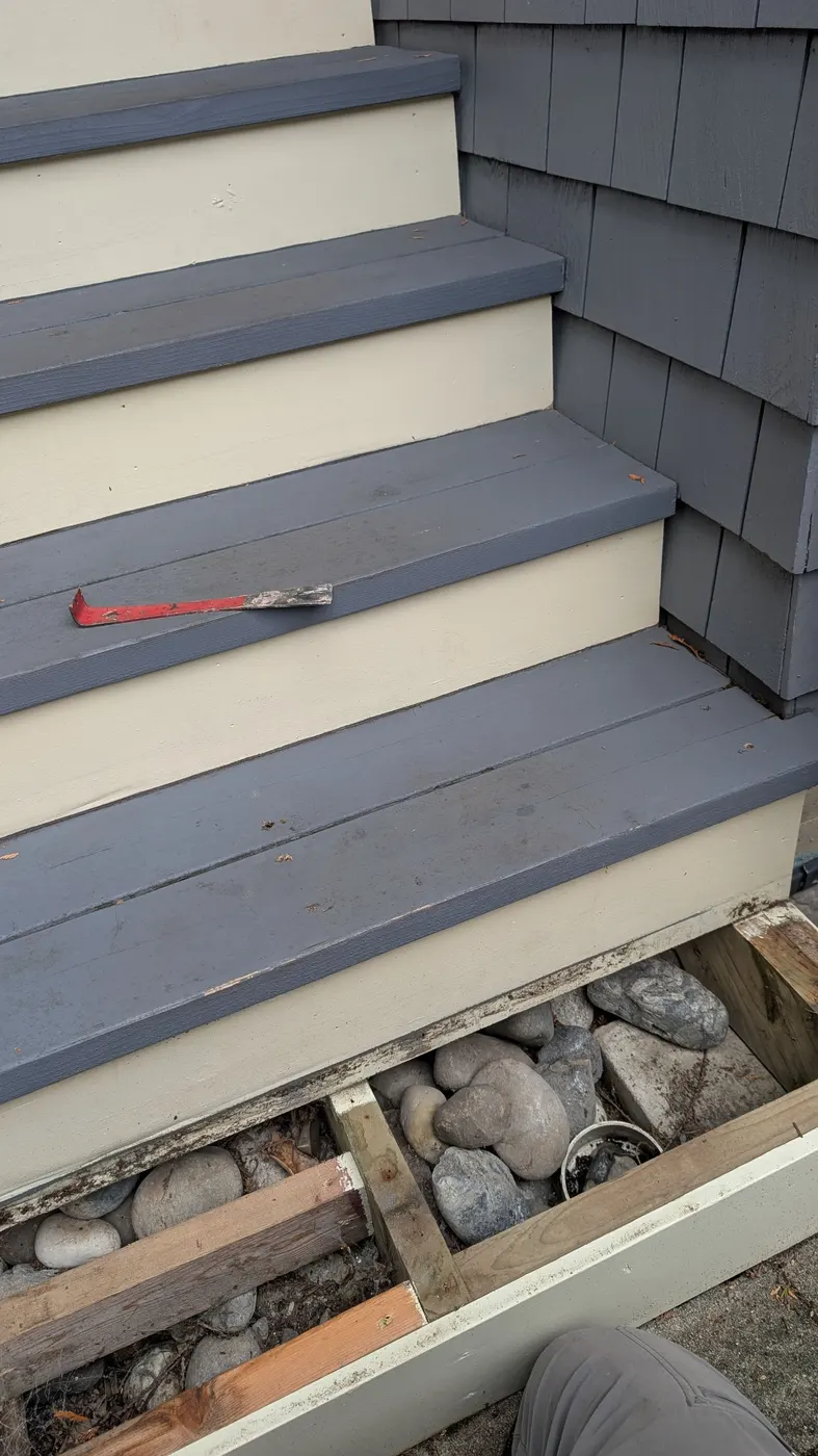 Exterior wooden stairs in Vancouver, BC with a pry bar on the tread and rotted framing exposed beneath the bottom step during demolition.
