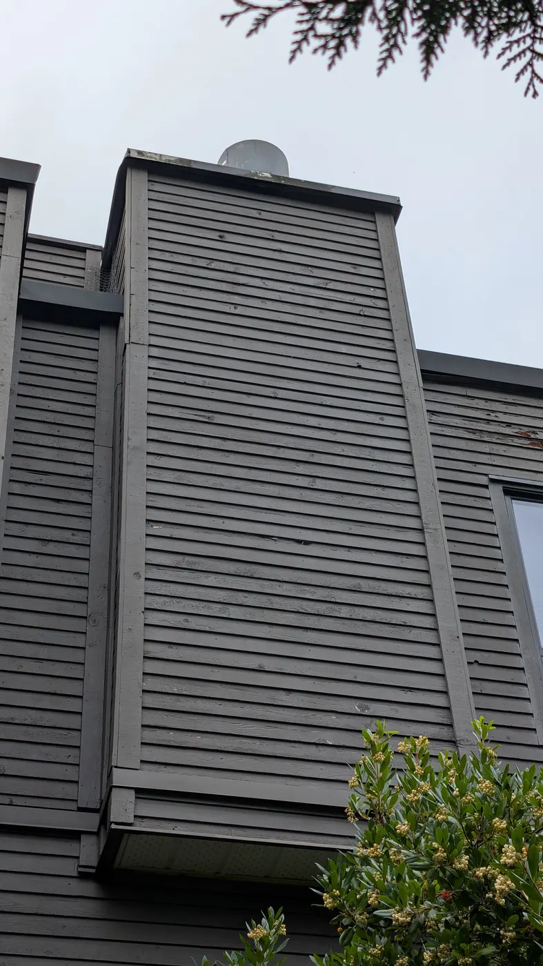 Weathered grey wood siding on a chimney chase at a Vancouver, BC strata building, viewed from below before carpentry repairs.