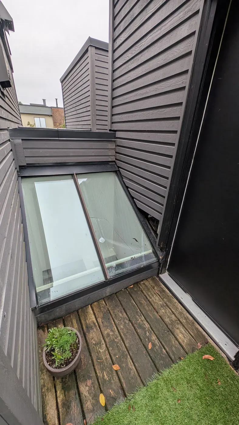 Wide view of a rooftop skylight window well and exterior siding at a Vancouver, BC townhome, showing the deck, potted plant, and cracked glass needing carpentry repair.