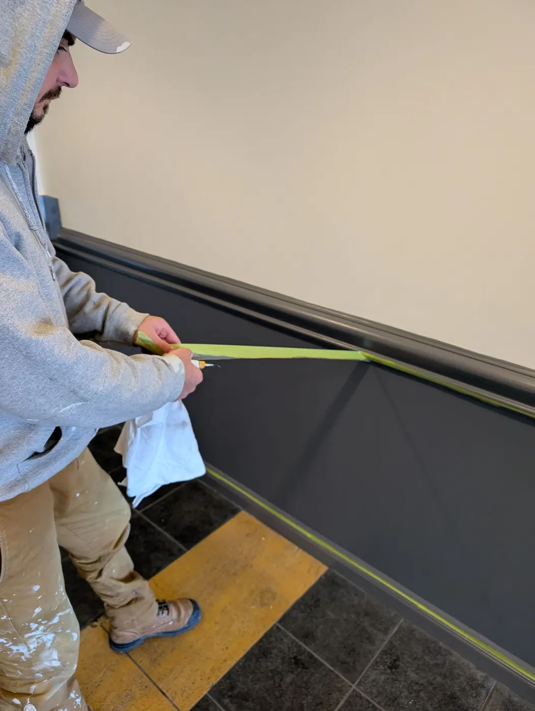 Painter pulling green painter's tape to reveal a crisp line between charcoal wainscoting and taupe walls in a Vancouver condo hallway.