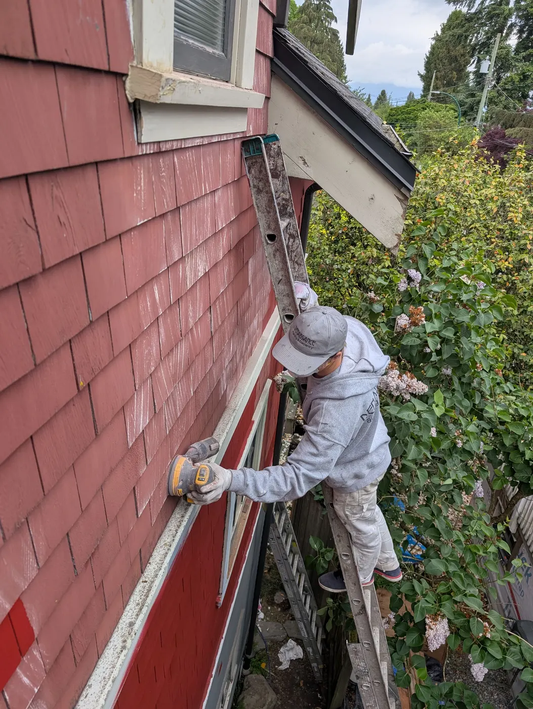 Renaissance Painting carpenter on a ladder using a drill to secure window trim on a Vancouver, BC craftsman home during exterior renovation.