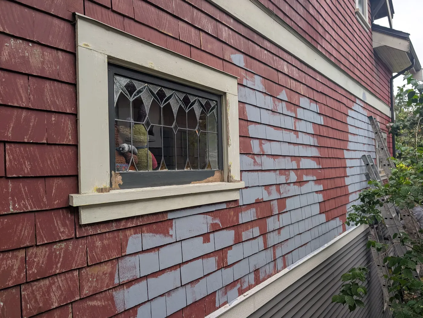 Exterior wall of a Vancouver, BC home mid-repaint, showing new blue-gray paint over the original red cedar shingles around a leaded glass window.