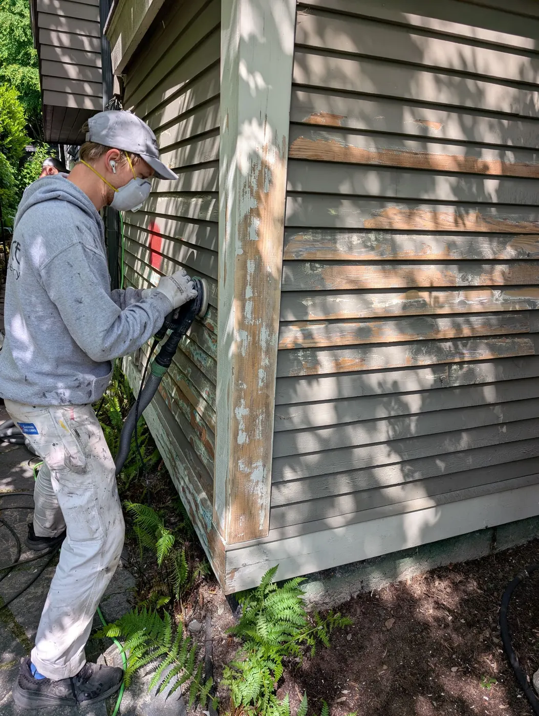 Painter sanding weathered wood siding down to bare wood with a dust-extraction sander on a Vancouver, BC home exterior.