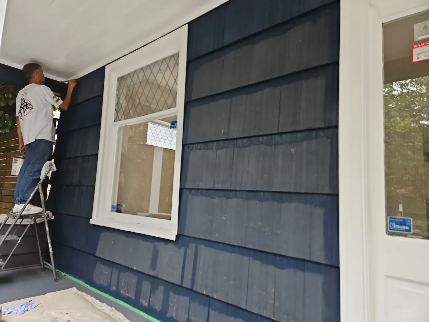 Painter on a ladder repainting navy shingle siding under a covered porch in Vancouver, BC.