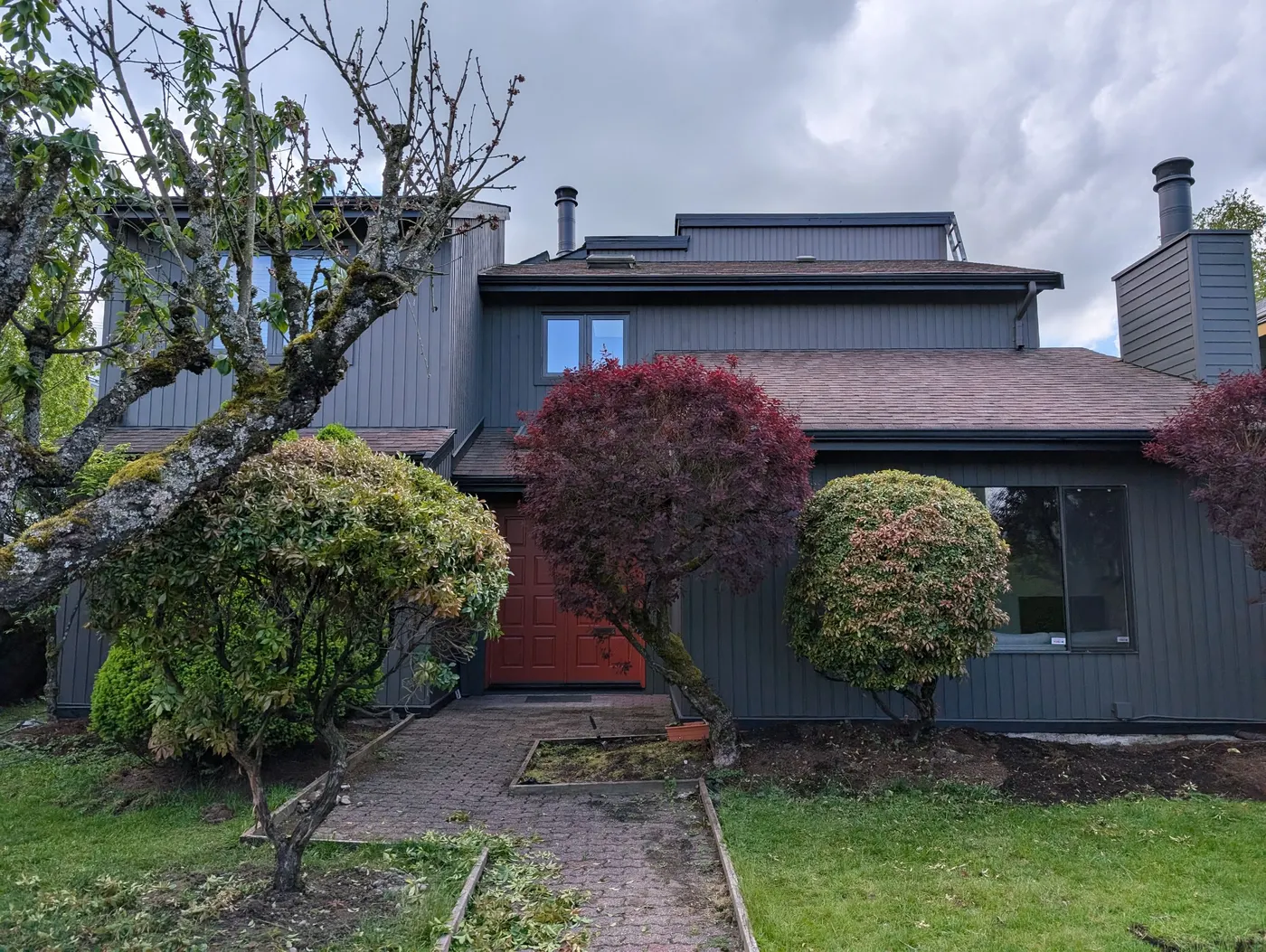 Completed dark charcoal gray exterior repaint with red front door on a Vancouver, BC home surrounded by landscaping.