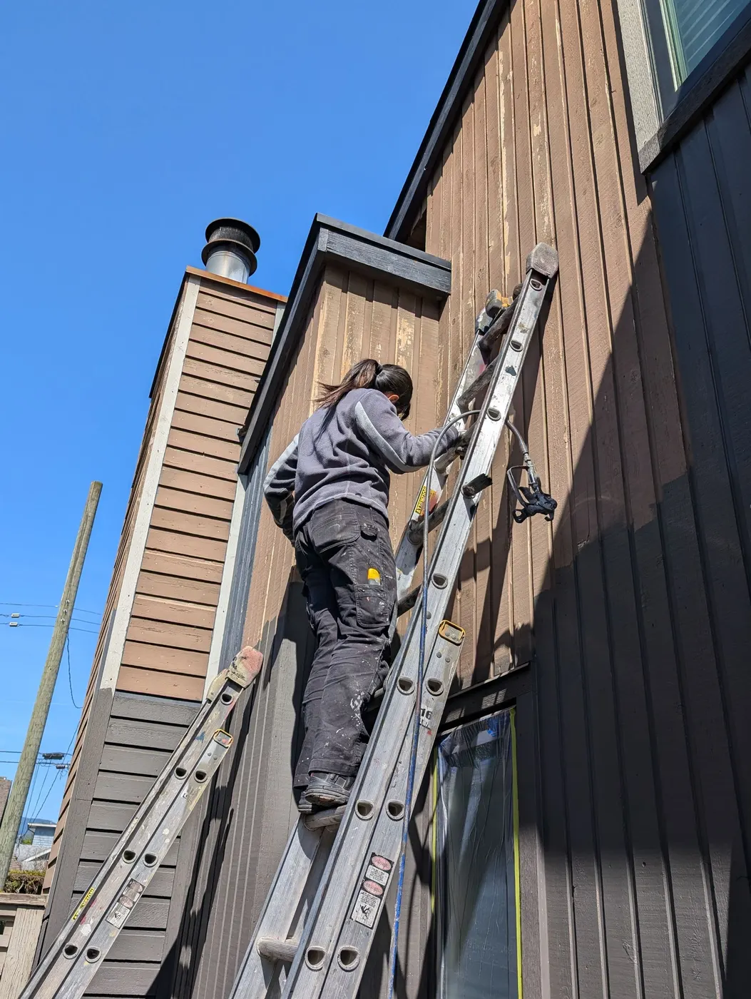 Renaissance Painting team member climbing a ladder to prep wood siding on a Vancouver, BC home under blue skies.