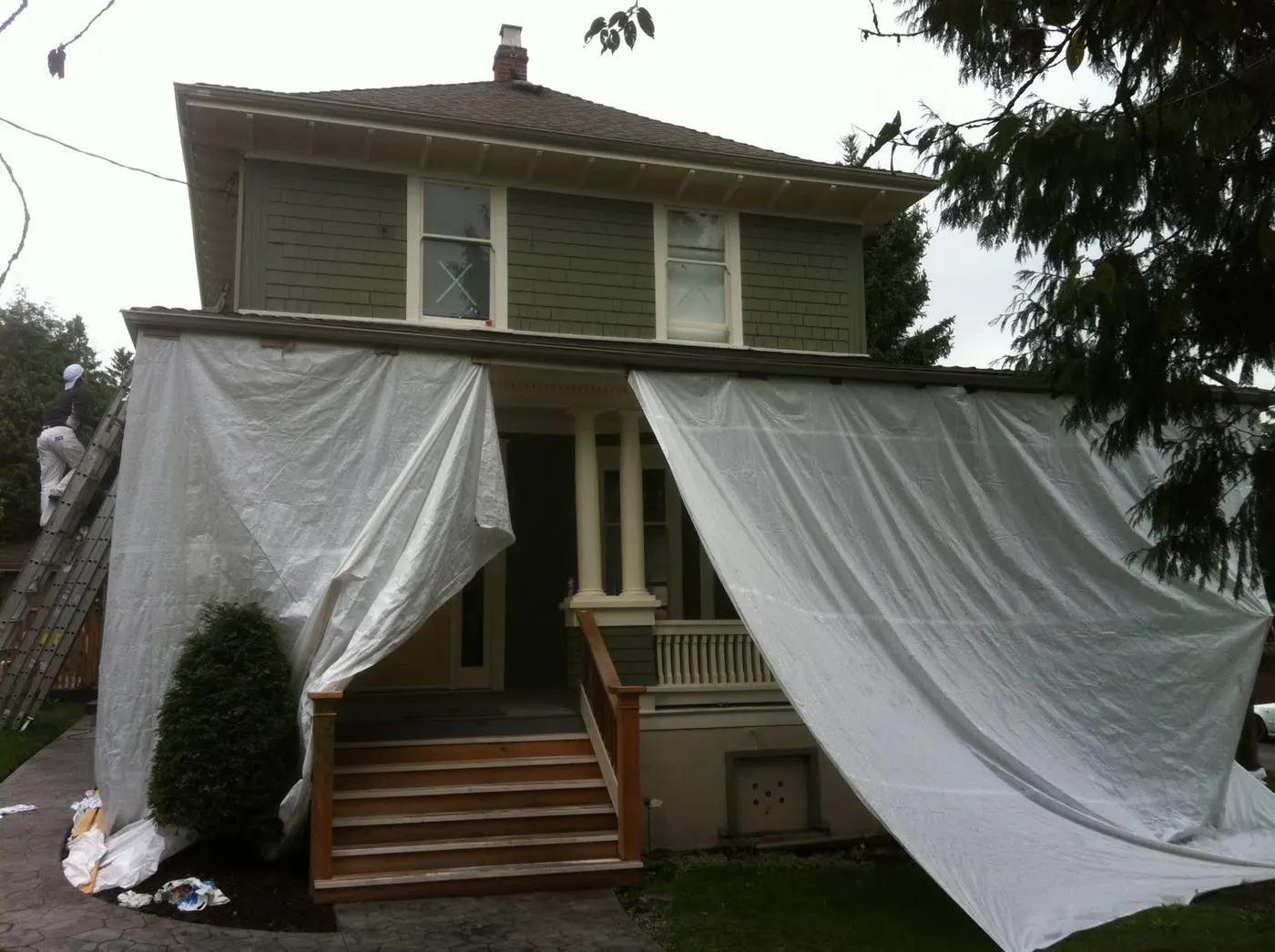 Painter on a ladder prepping a Vancouver character home for exterior painting, with protective drop cloths draped over the porch roof