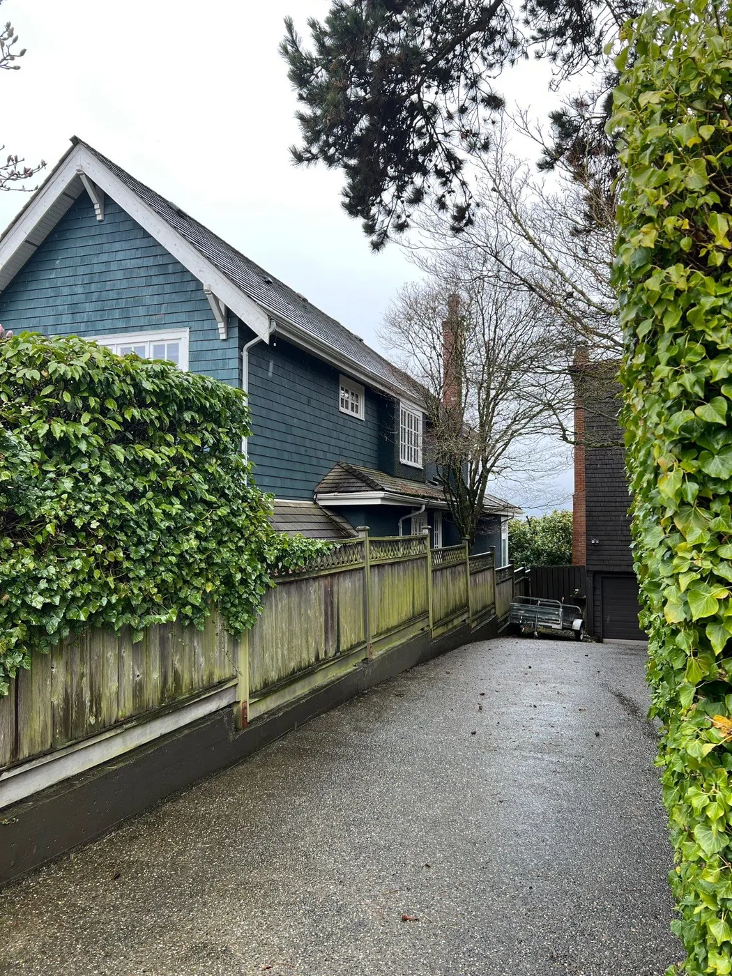 Driveway-side exterior of a blue-green character home in Vancouver, BC with wood fence and mature trees
