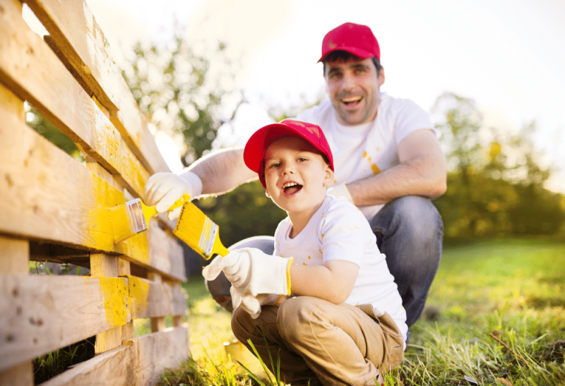 A father and a kid are both painting the fence in the yellow colour with brushes and having a good time.
