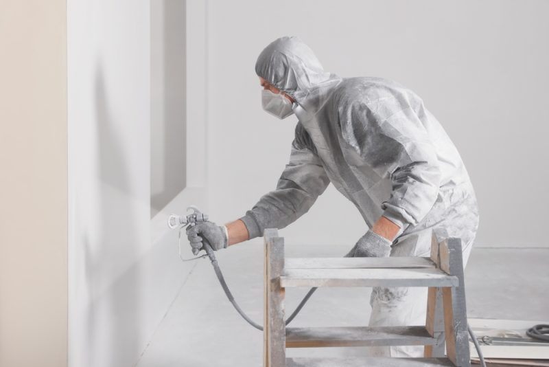 A man in a white protection suite is spar painting white room.