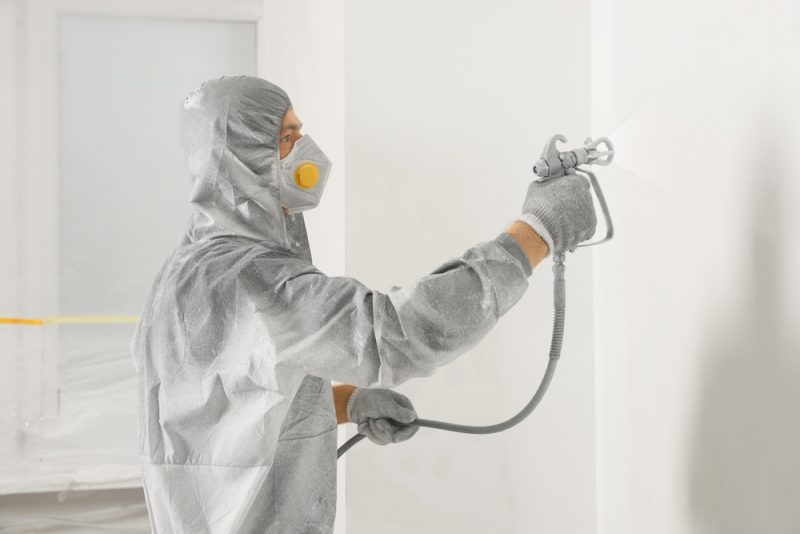 A spray painting professional in a disposable protective coverall suit is spray painting a wall in white colour.