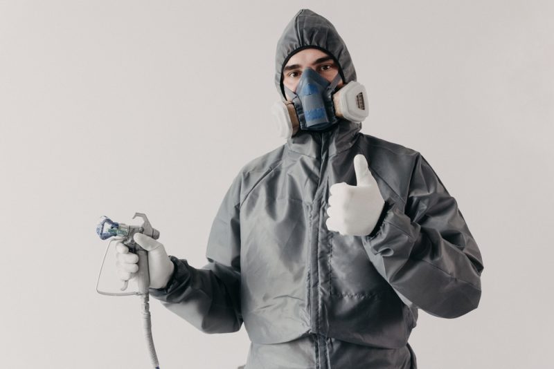 A painting contractor in a grey disposable protective coverall suit with a paint sprayer showing thumb up.