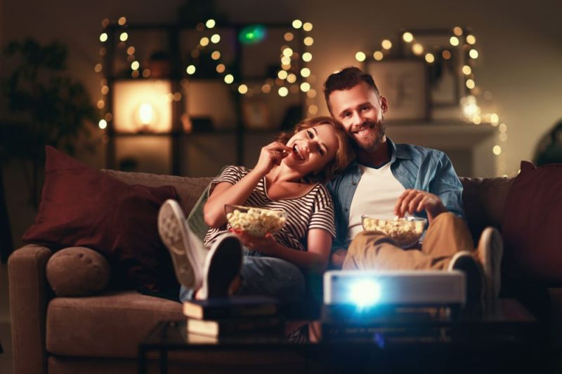 A happy couple is watching a movie on a projector while eating popcorn.