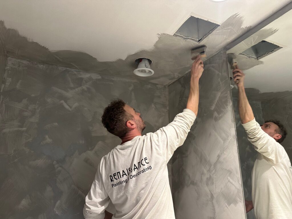 A painter is painting a celling in a grey colour in front of the mirror.