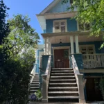 Front view of a blue Craftsman house with wide stairway, covered porch, and mature trees