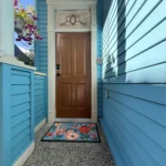 Front entry of a blue Craftsman house with wood door, decorative glass transom, and floral doormat