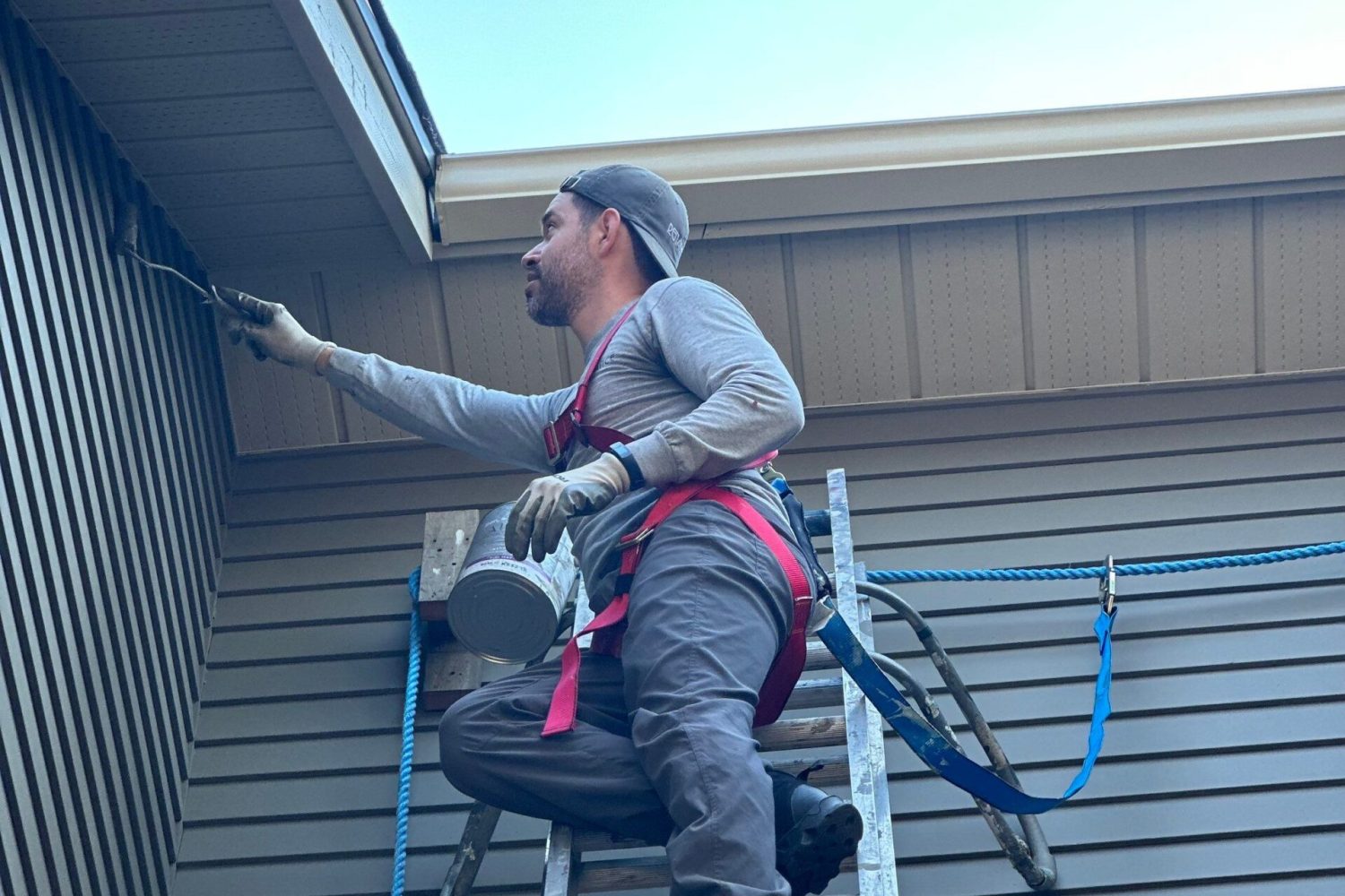 Painter on a ladder is painting a wooden siding a grey colour