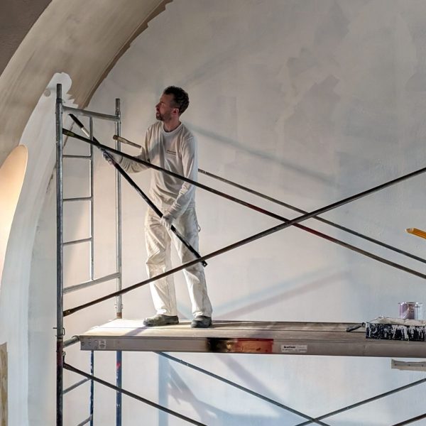 Painter on scaffolding applying white paint to a large curved interior wall with a roller at a commercial site.