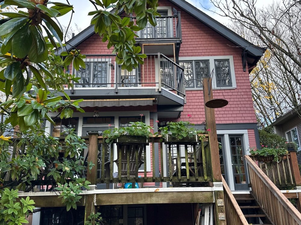 Back view of a three-storey Craftsman-style house with red shingle siding, wood deck, and surrounding autumn foliage."